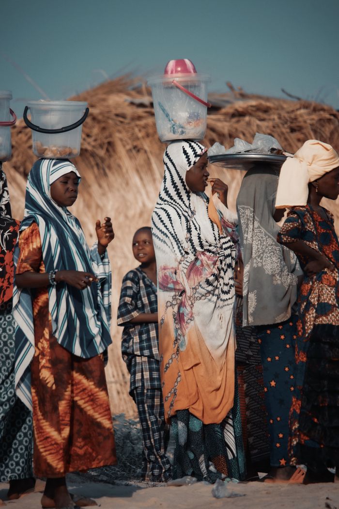 A group of African women standing outdoors in traditional clothing, some with baskets on their heads.